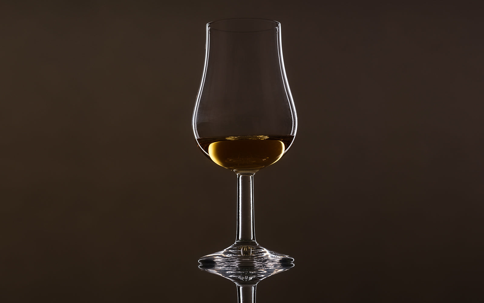 A stemmed tulip nosing glass of amber whisky side-lit on a dark background, the vessel for the discipline of nosing. Baroque Whisky Australian Single Malt.
